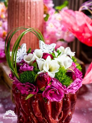 Composition florale dans un vase rouge, fleurs blanches et violettes, papillons décoratifs, fond rose.